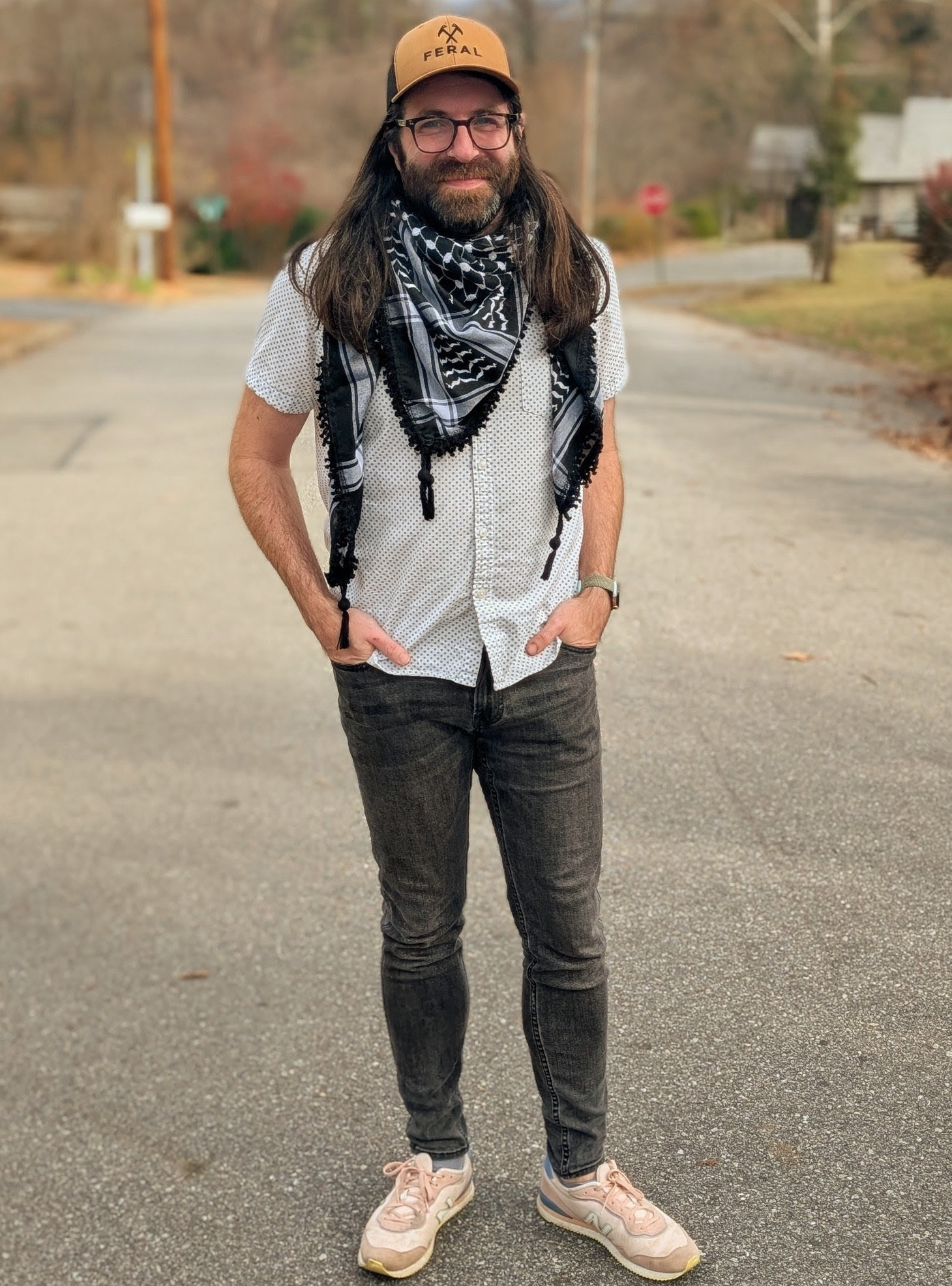 Dan Ferrell, a non-binary person with glasses, long brown hair and a beard, with a white and black keffiyah and glasses, smiling on a stree.